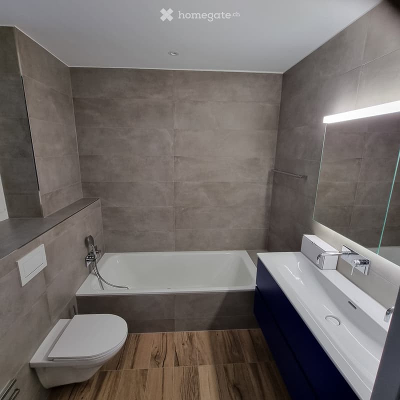 Modern bathroom with a bathtub, toilet, sink, and large mirror. Features grey tiled walls and wooden flooring.