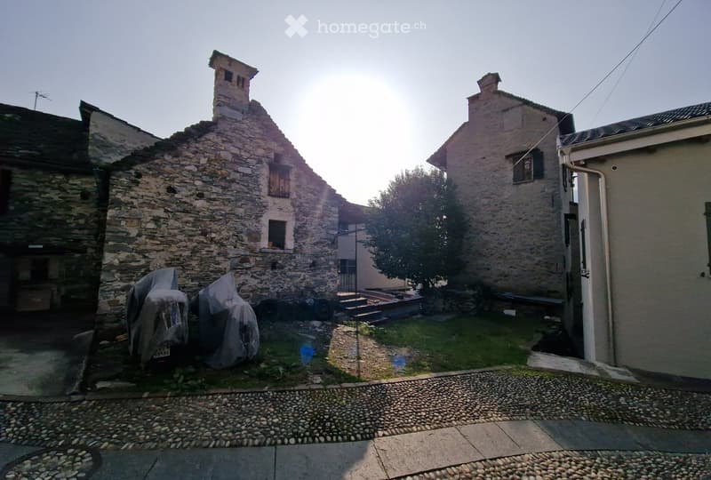A rustic stone building with a small chimney, partially covered in ivy, surrounded by a small grassy area and a covered vehicle.