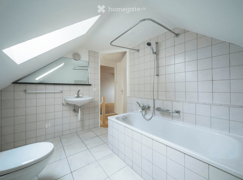 Modern bathroom with white tiles, a bathtub, toilet, sink, mirror, and a shower area with a glass enclosure. The room features a skylight providing natural light.