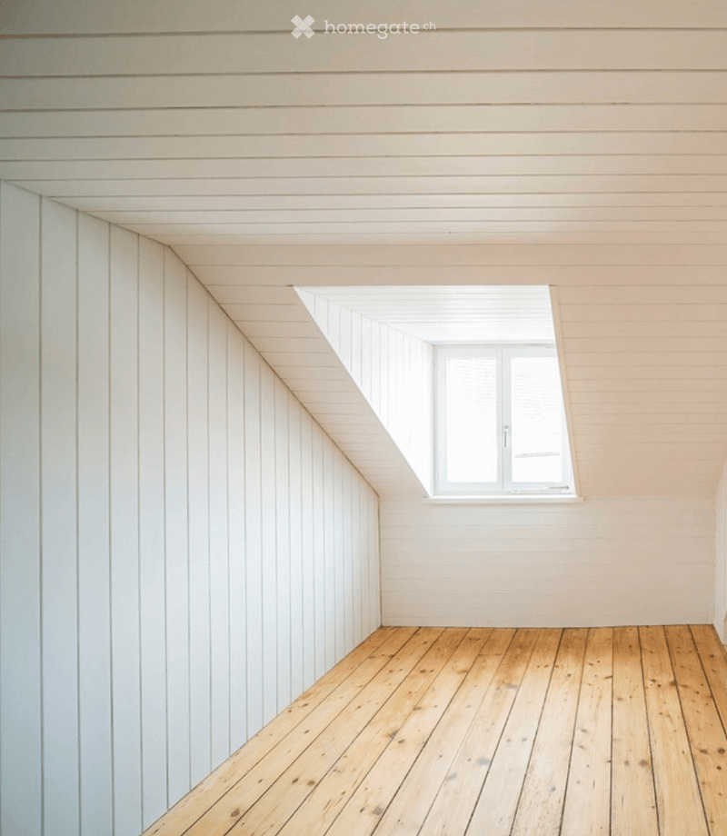 Empty room with wooden flooring and white paneled walls, featuring a slanted ceiling and a single window with white frame.