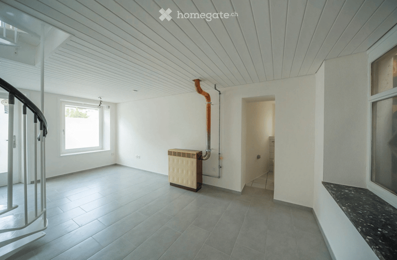 Empty room with white walls, tiled floor, and a curved staircase leading upwards. A radiator is visible against one wall, and a doorway leads to another space.