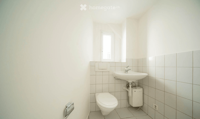 A small, clean bathroom with white tiled walls and floor, featuring a toilet, a sink with a faucet, and a toilet paper holder.
