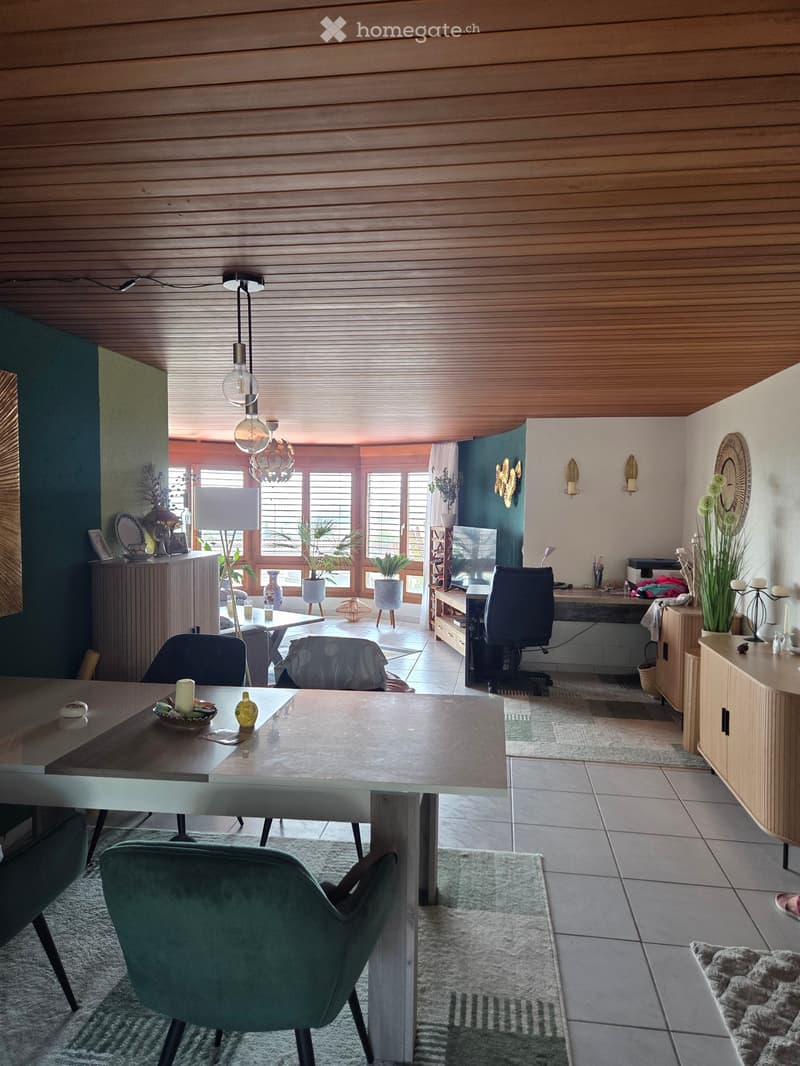 A modern, open-concept living and dining area with a wooden ceiling, large windows, and a mix of contemporary and rustic furniture. The space includes a dining table with chairs, a cabinet, a desk with an office chair, and decorative items such as lamps,