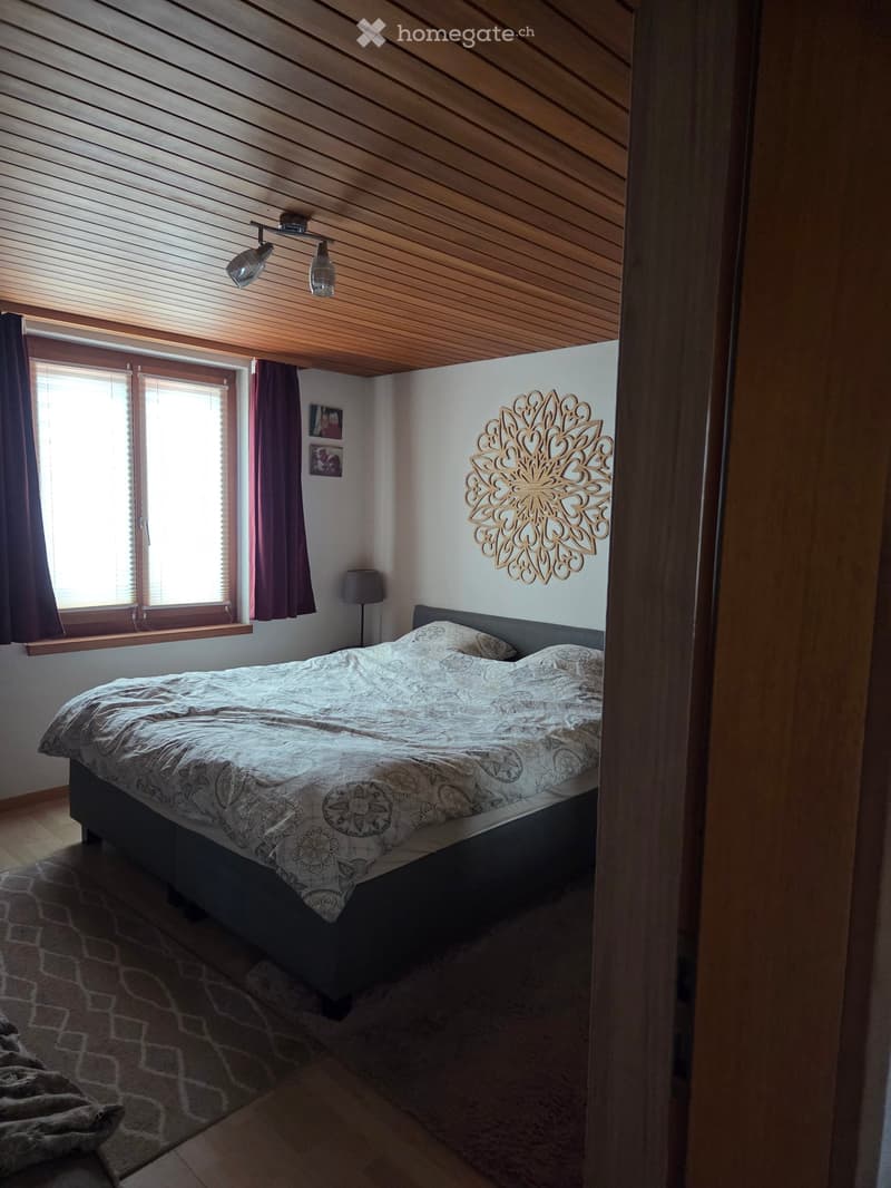 Cozy bedroom with a wooden slanted ceiling, a large window with dark curtains, a bed with patterned bedding, a wall-mounted decorative piece, and a bedside lamp.