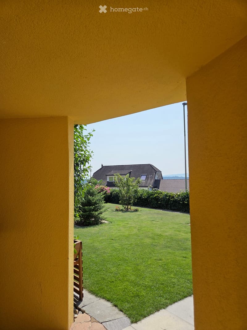View from a covered patio onto a well-maintained lawn with a house and garden in the background.