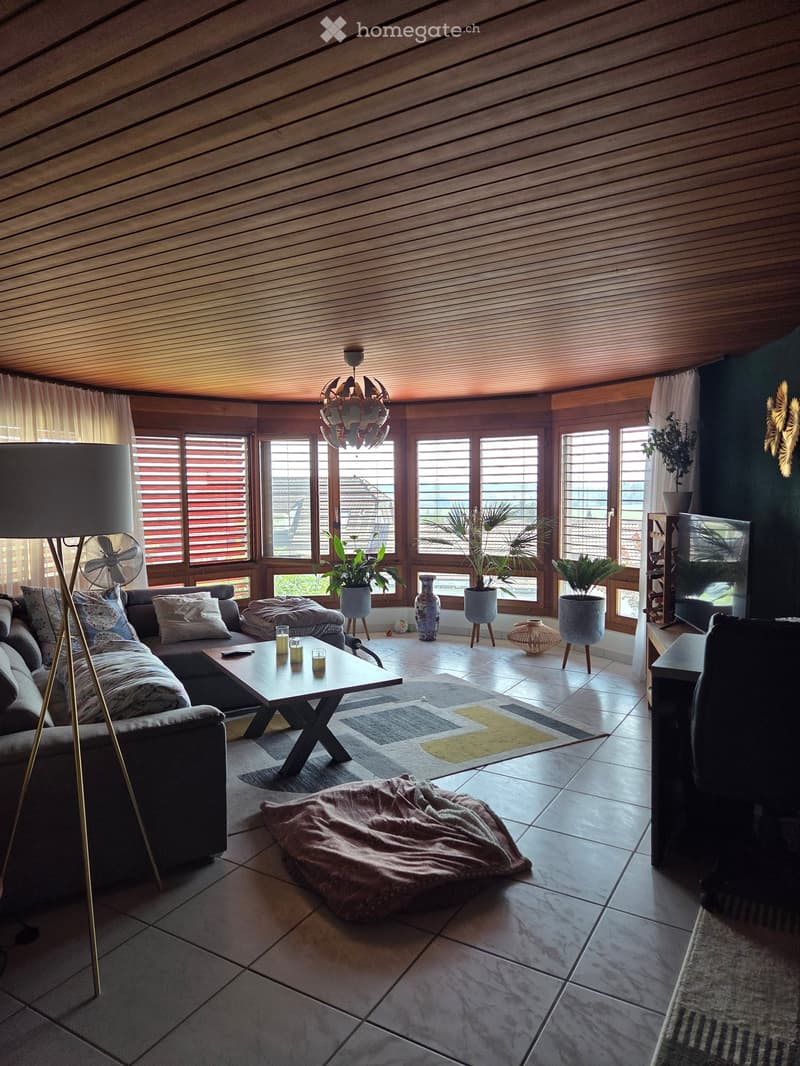 Modern living room with large windows, wooden ceiling, grey sofa, coffee table, floor lamp, fan, and decorative vases.