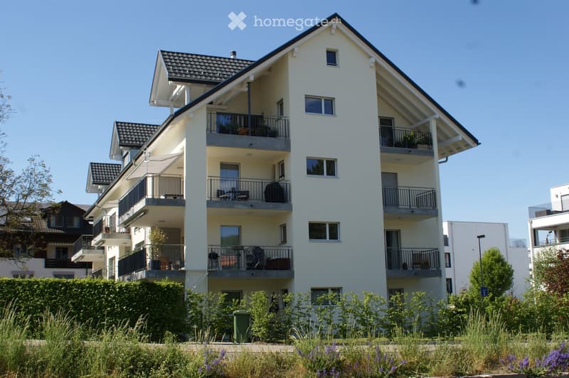 Modern multi-story residential building with balconies, large windows, and a well-maintained garden in front.