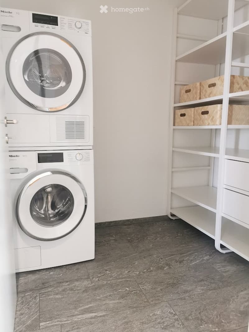 A modern laundry room with a stacked washer and dryer unit, a white shelving unit with storage boxes, and a dark grey tiled floor.