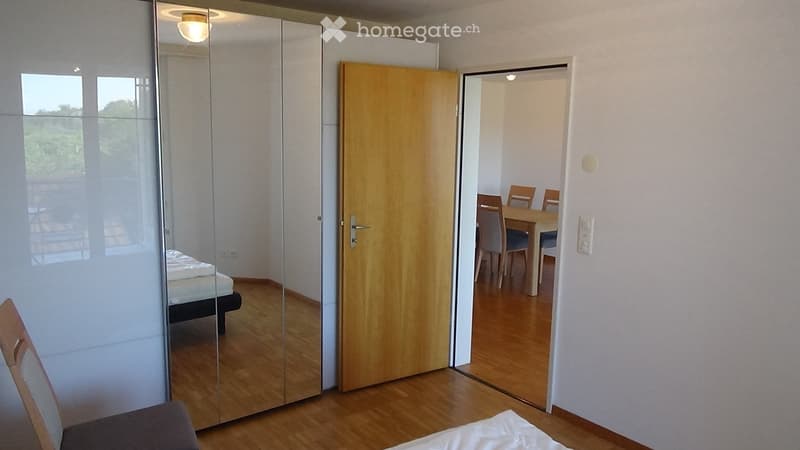 The image shows a furnished room with a bed, a large mirror, a wooden door, and a view into an adjacent dining area with a table and chairs.