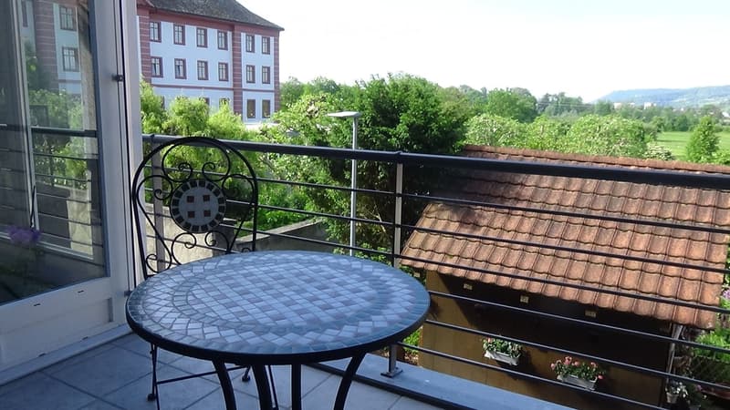 Balcony with a round mosaic table and a decorative metal chair, overlooking a green landscape and a multi-story building.