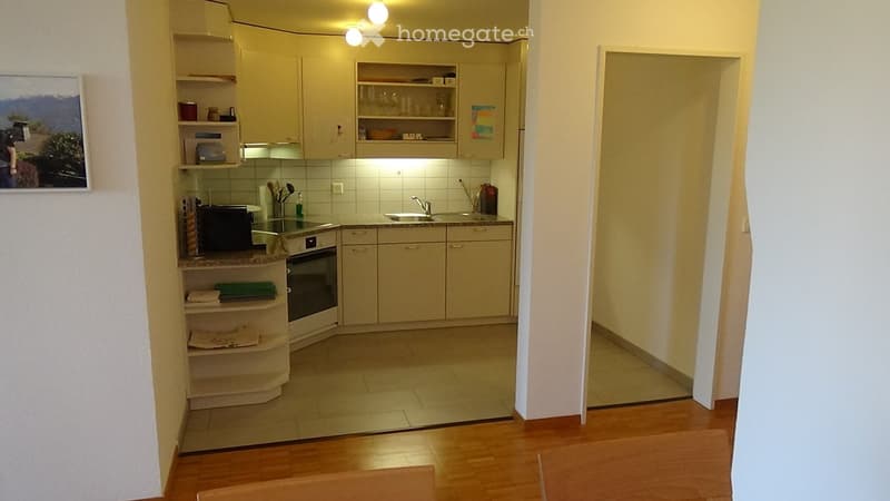 The kitchen features light-colored cabinets, a tiled backsplash, and a granite countertop. Visible appliances include an oven, microwave, and sink. Shelves hold various kitchen items, and a doorway leads to another room with wooden flooring.