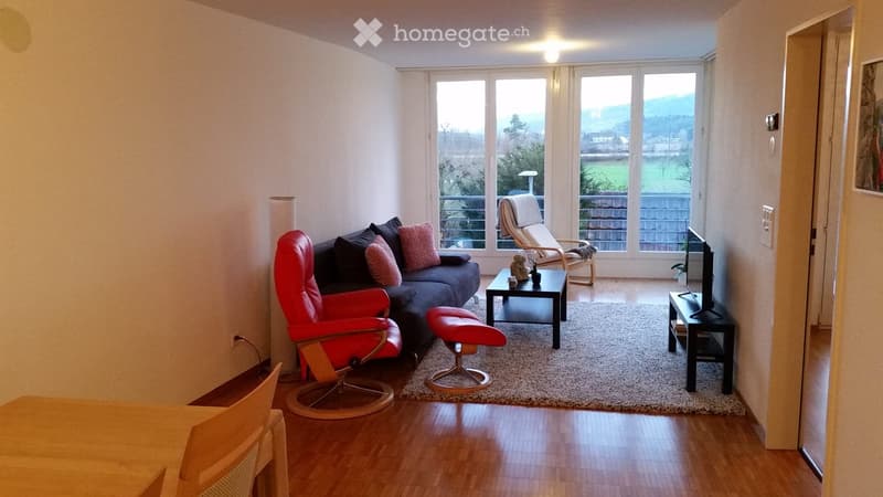 Modern living room with large windows offering a scenic view, featuring a red sofa, coffee table, red chair, ottoman, and television stand.