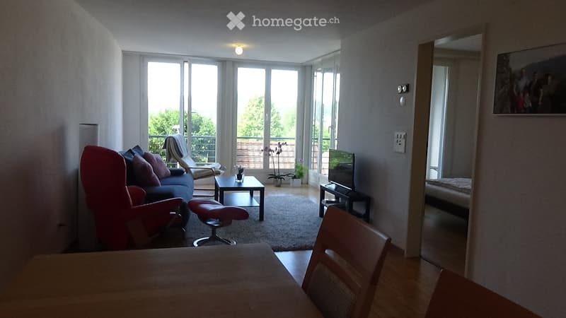 Bright living room with large windows and a sliding glass door leading to a balcony. Features a red armchair, blue sofa with pillows, a coffee table, a TV stand, and a dining table with chairs.