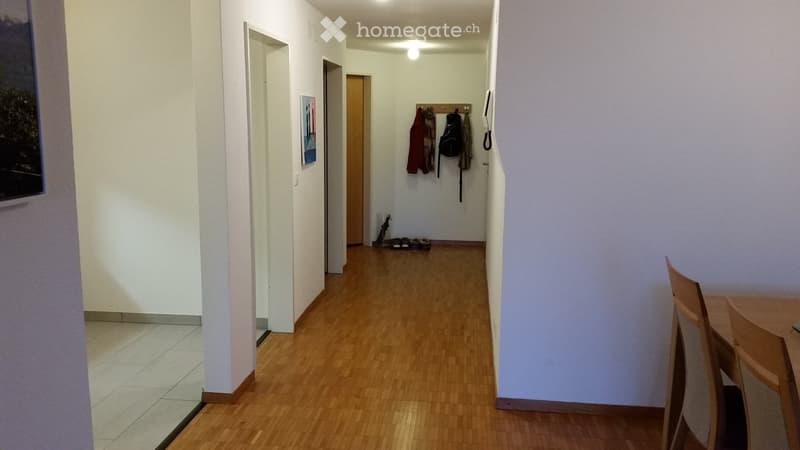 A narrow hallway with wooden flooring, white walls, and several doors leading to different rooms. A coat rack with jackets and a backpack is visible, along with a small table and chairs in the foreground.