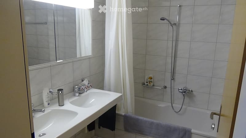 Modern bathroom with double sink, mirror, bathtub, and showerhead. Grey tiles and white fixtures.