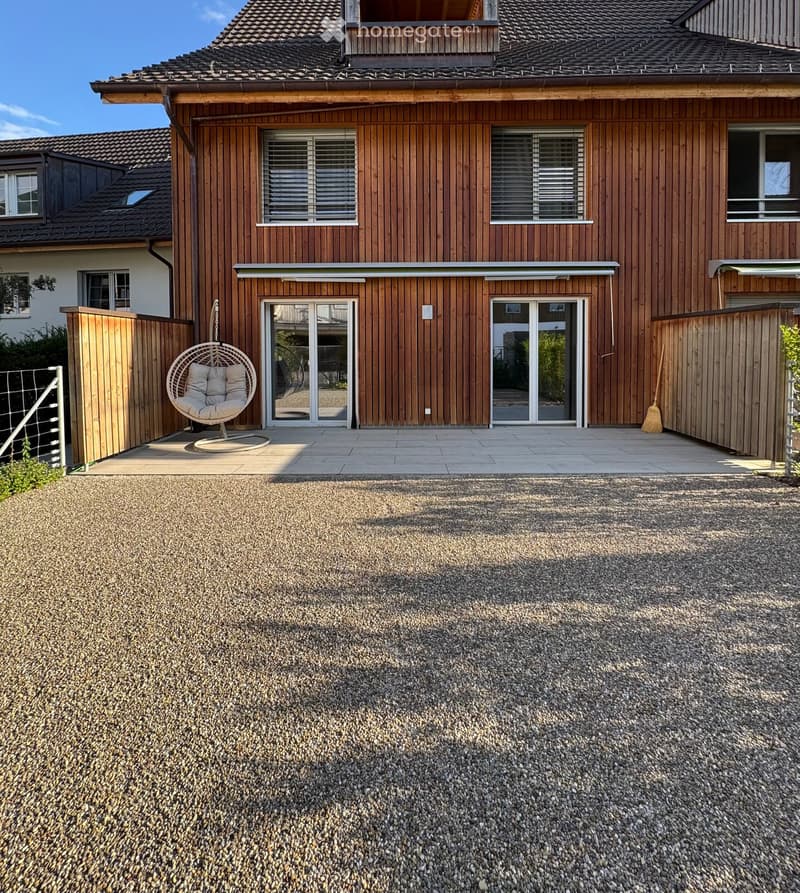Modern wooden house with large windows and sliding glass doors, featuring a gravel driveway and a wicker hanging chair on the patio.
