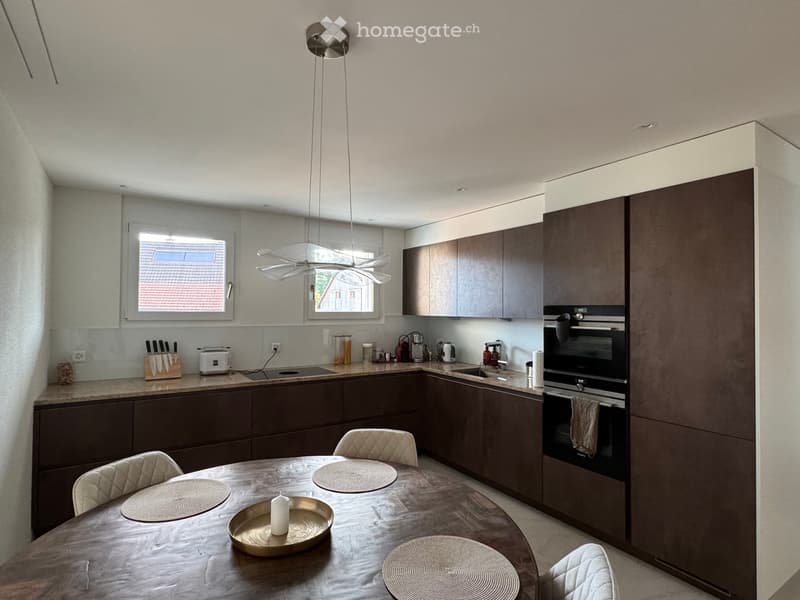 Modern kitchen with dark brown cabinets, a central dining table with four chairs, and various appliances including an oven, fridge, and toaster. The room features a sleek pendant light fixture and a window offering a view of the roof.