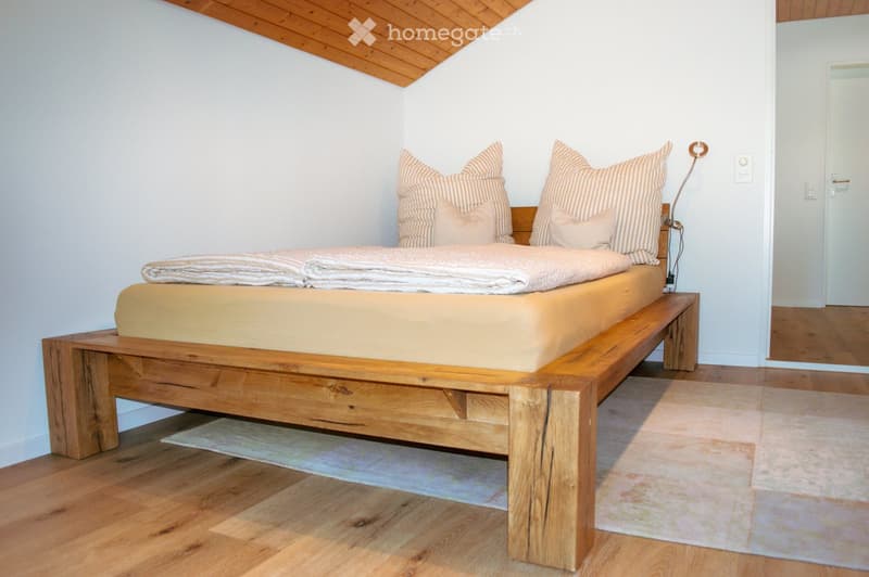 Cozy bedroom with a wooden bed, striped pillows, and a warm-toned blanket. The room features wooden flooring and a slanted wooden ceiling.