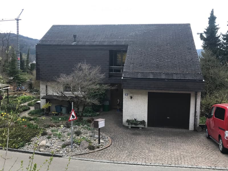 Two-story house with dark wooden exterior, garage, and landscaped garden with flowers and shrubs.