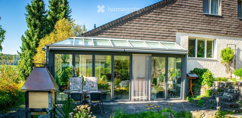 Outdoor patio with a glass-enclosed area, dining table and chairs, surrounded by greenery and a barbecue.