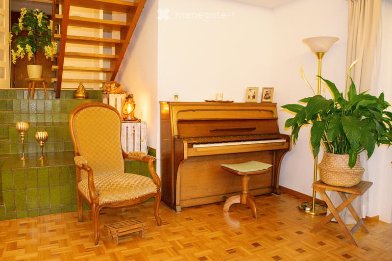 A well-furnished room with a wooden floor, a grand piano, a chair, a small table, a floor lamp, and decorative plants.
