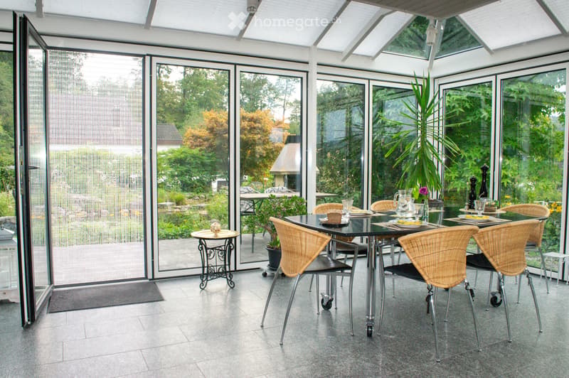 A spacious, well-lit conservatory with large glass walls and a retractable roof, featuring a dining area with a black table, wicker chairs, and various decorative items.