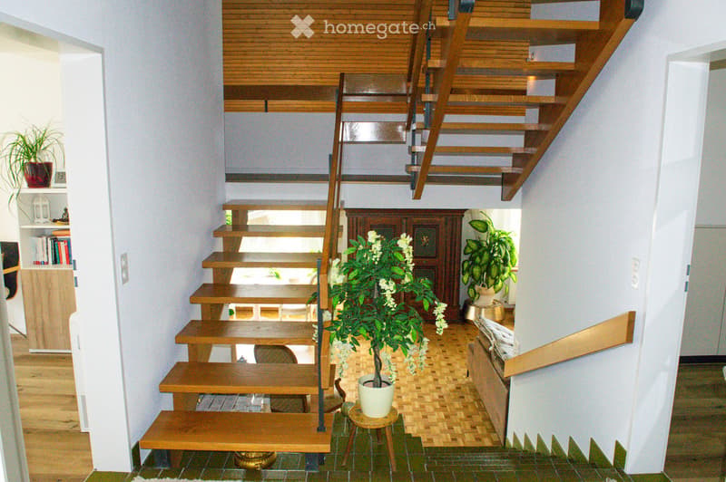 A modern hallway with a wooden staircase, potted plants, and a small table with a plant in the center. The walls are white, and the flooring is a mix of wood and tile.