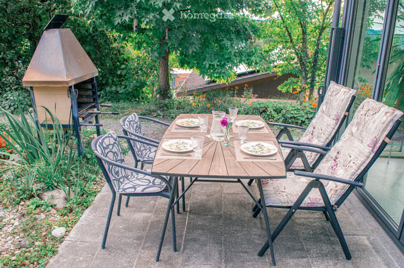 Outdoor patio area with a wooden dining table, four chairs, plates, glasses, and a vase with a flower. A barbecue grill is visible to the left, surrounded by greenery and plants.