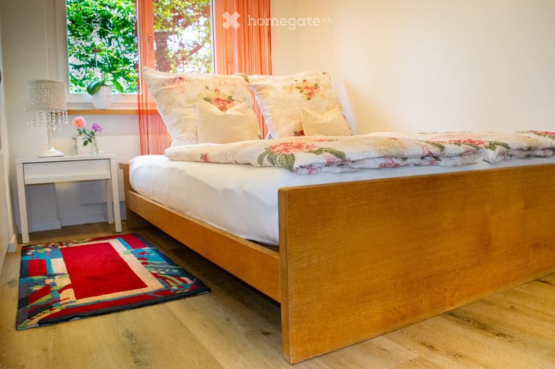 Cozy bedroom with a wooden bed, floral bedding, a nightstand with a lamp, and a colorful rug on the floor.
