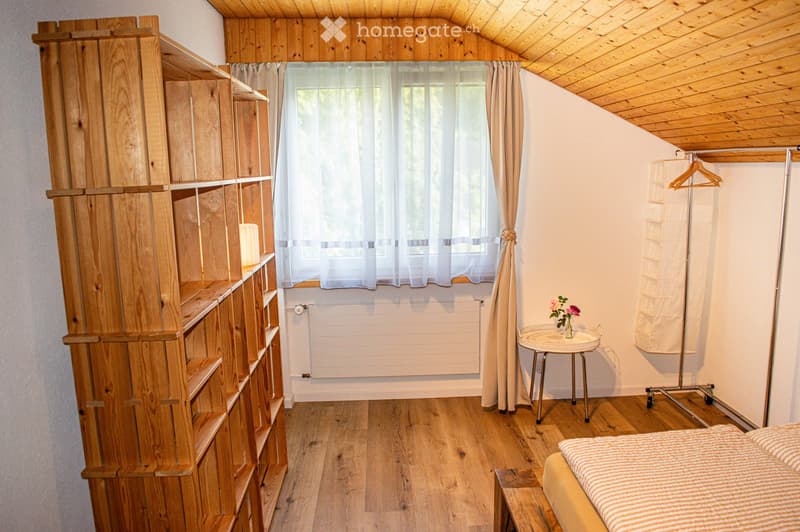 Cozy bedroom with wooden walls and ceiling, featuring a bed, wooden shelf, small table with flowers, and a window with curtains.