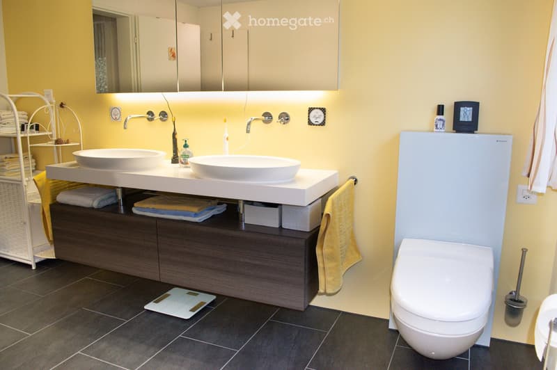 Modern bathroom with double sink vanity, dark wood cabinet, white ceramic sinks, large mirror, and a toilet. The space features yellow walls, dark tiled flooring, and various bathroom accessories including towels, a scale, and toiletries.