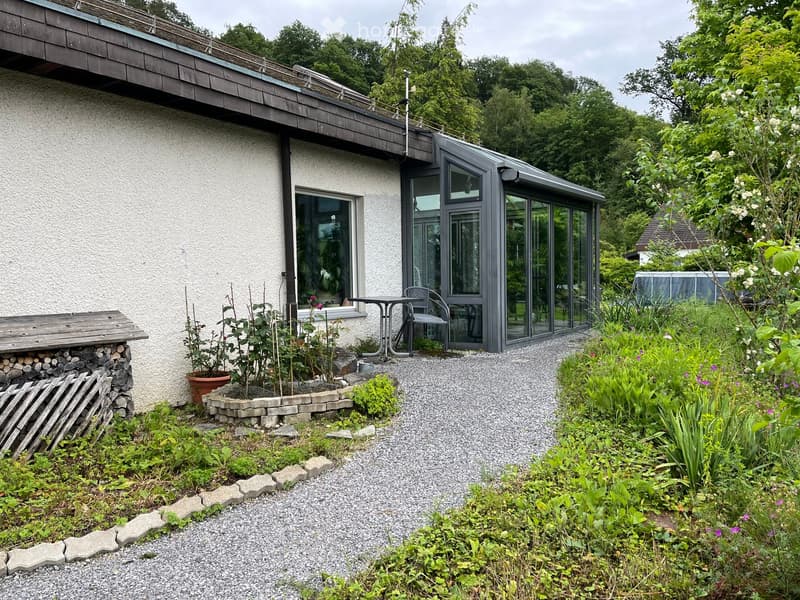 Garden area with a gravel pathway, wooden bench, potted plants, and a small table and chair near the house entrance.