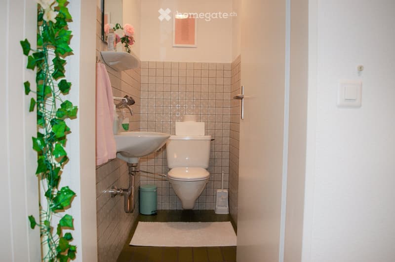 A compact bathroom with a white sink, toilet, and a pink towel hanging on the wall. The walls are tiled with a light gray pattern, and a small white rug is placed on the floor. A flower vase with pink flowers sits on the sink, and a mirror is mounted abov