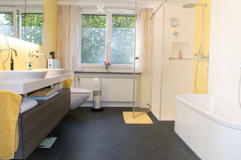 Modern bathroom with double sink, toilet, bathtub, shower, and large window with frosted glass. Features dark tiled floor, light yellow walls, and a wooden cabinet with towels.