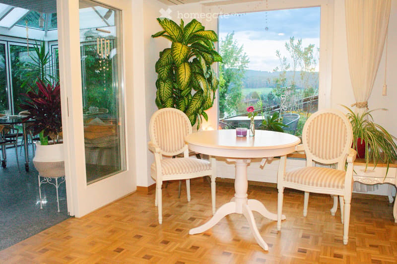 A well-lit room with wooden flooring, large windows, and a round white table with two matching chairs. A vase with a flower and a candle are placed on the table. Potted plants are visible near the windows, and the room has a cozy, elegant ambiance.