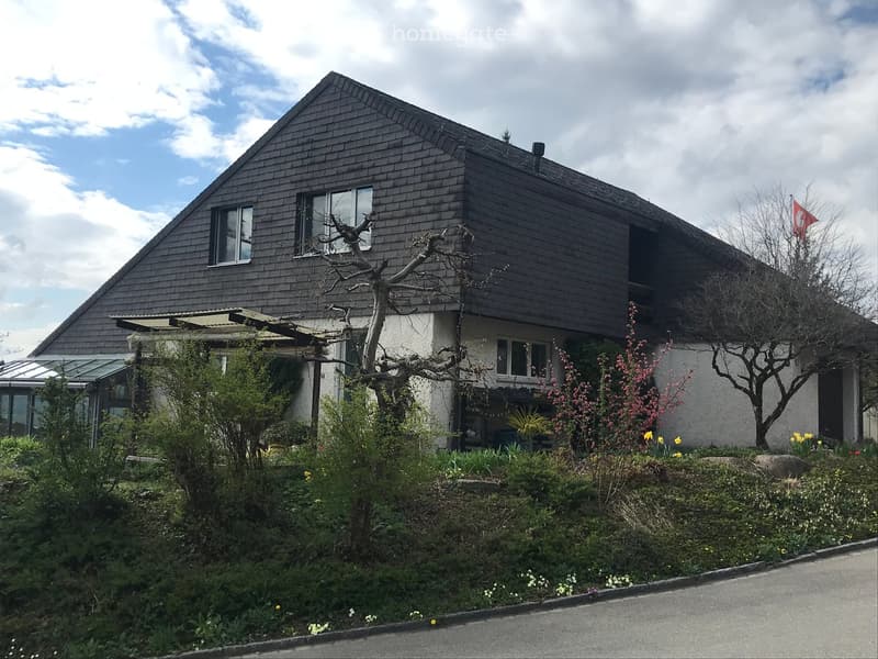 Two-story house with dark wooden exterior, multiple windows, and a well-maintained garden with flowering plants and trees.