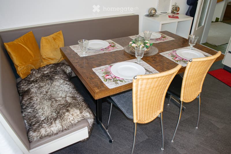 Cozy kitchen with a wooden dining table set for four, complete with place settings, a floral table runner, and a bench with a fur throw and yellow pillows.
