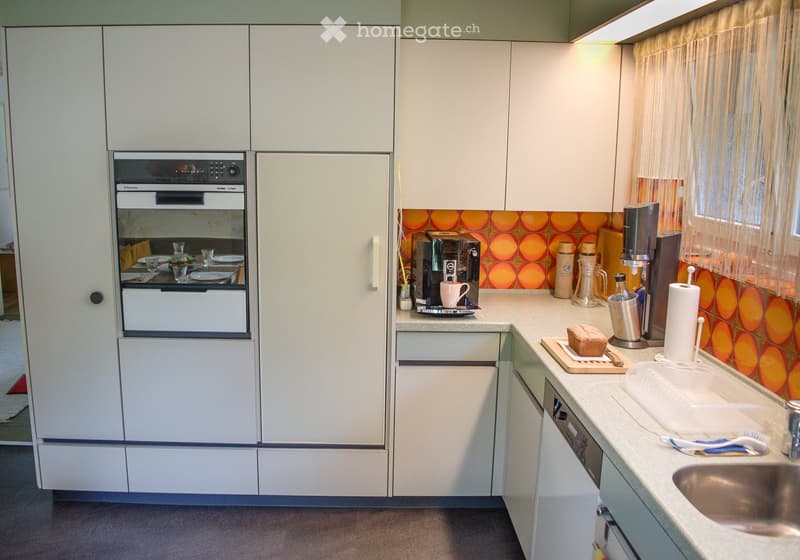 Modern kitchen with white cabinets, stainless steel appliances, and a colorful backsplash. Features an oven, coffee maker, sink, and various small items on the counter.