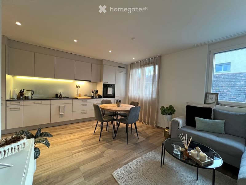 Open-plan living and dining area with modern furniture, including a sofa, coffee table, dining table, and chairs. The kitchen features light-colored cabinets, an oven, and a countertop with various items. The space is well-lit with recessed lighting and h