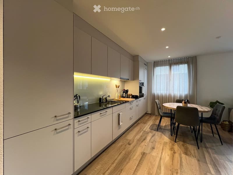 Modern kitchen with white cabinets, wooden flooring, and a small dining area with a table and chairs. The kitchen features a sink, faucet, oven, and coffee maker, with a window covered by a curtain and a small plant in the corner.