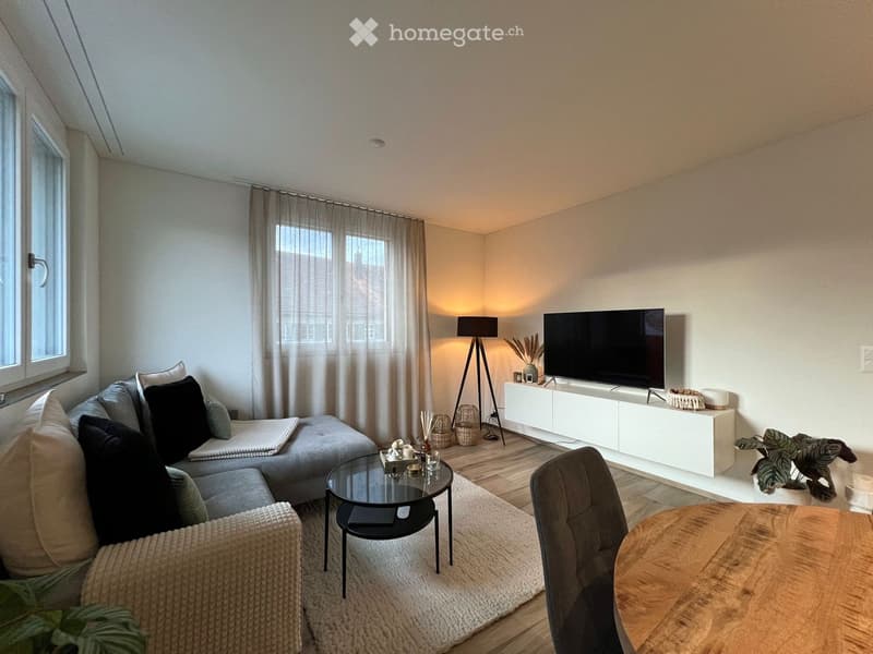 Modern living room with a grey sectional sofa, coffee table, TV stand, floor lamp, and a wooden dining table.