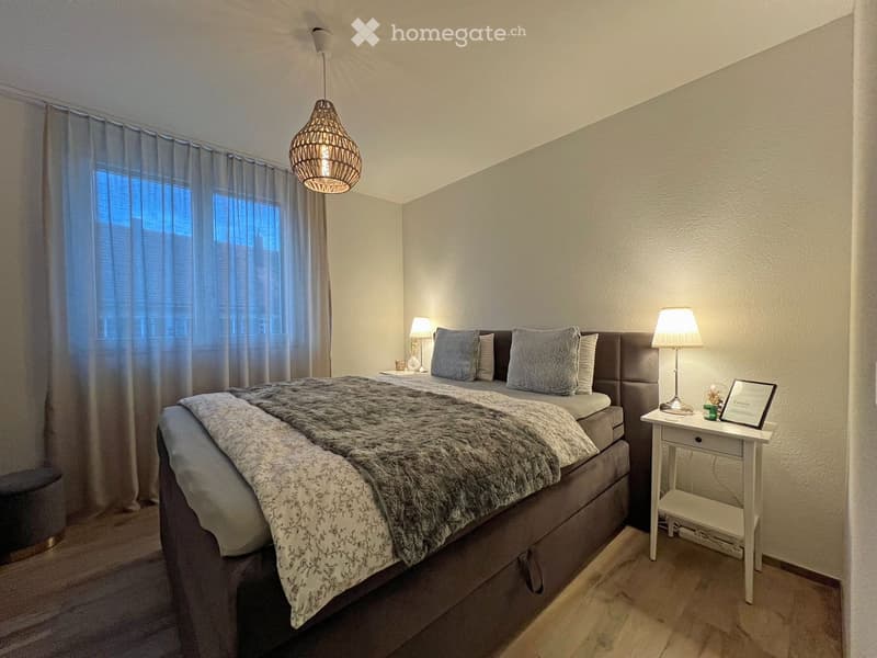 Cozy bedroom with a large bed, nightstands, lamps, and a hanging pendant light. The room features light-colored walls, wooden flooring, and sheer curtains on the windows.