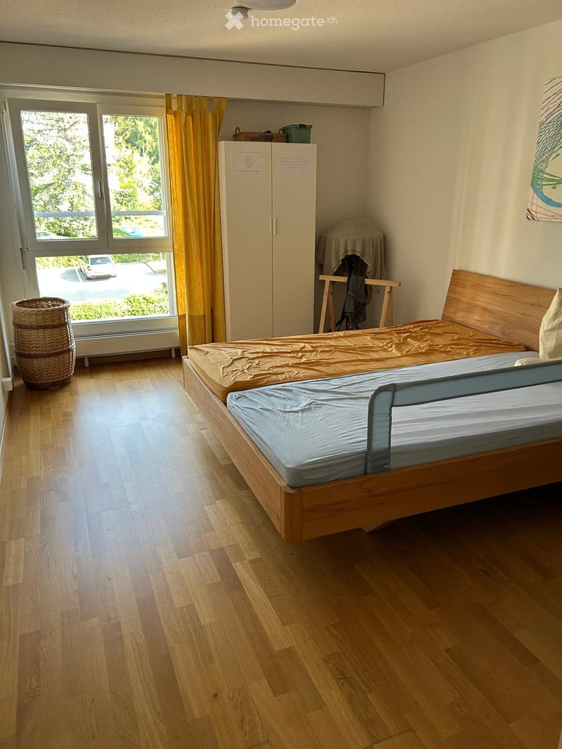A small bedroom with wooden flooring, a single bed with a wooden headboard, a wardrobe, and a wicker basket near the window.