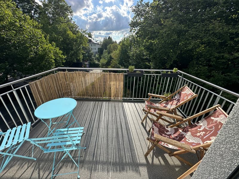 Balcony with a turquoise table, two turquoise chairs, and two wooden deck chairs with patterned cushions, surrounded by a metal railing and bamboo screen.