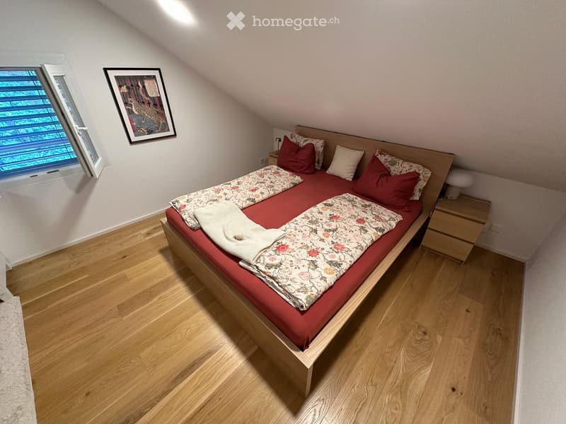 Cozy bedroom with wooden flooring, a double bed with red bedding and floral duvet, nightstand, and a framed picture on the wall.
