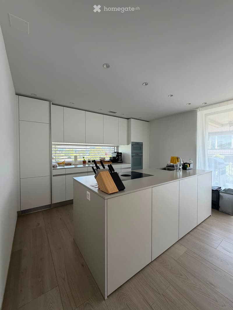 Modern kitchen with white cabinets, a central island, and stainless steel appliances. The island features a knife block and a sink with a modern faucet. The countertops are light-colored, and the floor is made of light wood.
