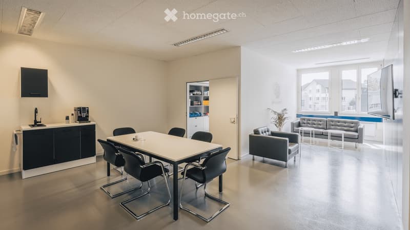 Modern office break room with a kitchen area and lounge seating. Features a white table with black chairs, a grey sofa, a small coffee machine, and a sink with a faucet.