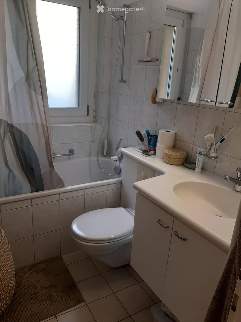 Bathroom with a toilet, sink, bathtub, mirror, and various toiletries on the sink counter. The room features tiled walls and a window with a shower curtain.