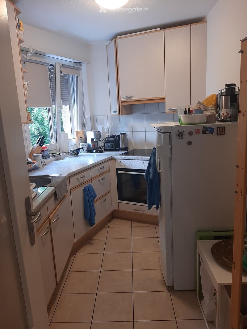 Compact kitchen with white cabinets, tiled floor, and various appliances including a refrigerator, oven, coffee maker, and blender. A window with blinds allows natural light to enter.