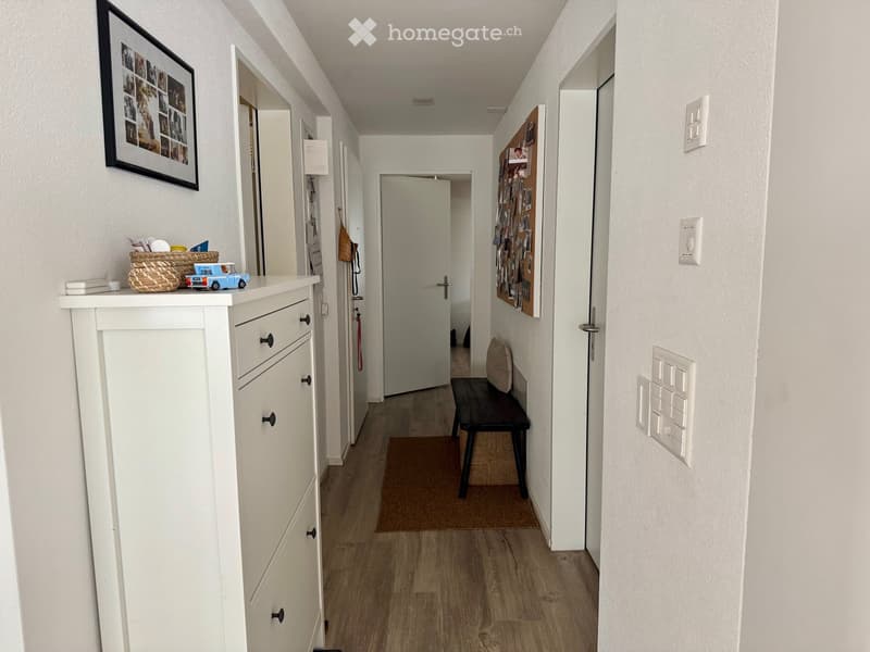 A narrow hallway with light wooden flooring, white walls, and several doors leading to other rooms. A small bench with a cushion sits near the end, and a white cabinet with drawers is positioned on the left side, topped with a woven basket and small decor